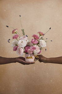 Two hands holding a bouquet of flowers with 'MOM' written on a jar against a beige background