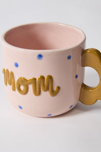 Pink mug with 'mom' in gold and blue polka dots on a white background