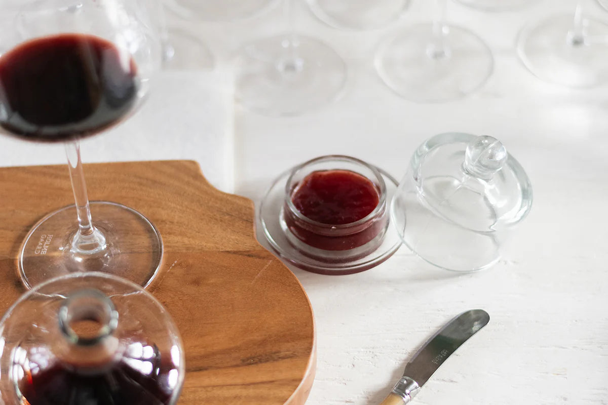 Red wine in a glass with a wooden cutting board and a small container of jam on a white surface.