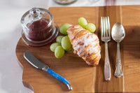 Wooden board with croissant, grapes, jam jar, knife, fork, and spoon on a white surface