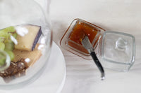 Square jar of jam with a knife next to a dessert with grapes and cheese on a white surface