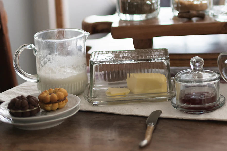 Butter dish with a knife, jar of jam, and pitcher of milk on a wooden table.