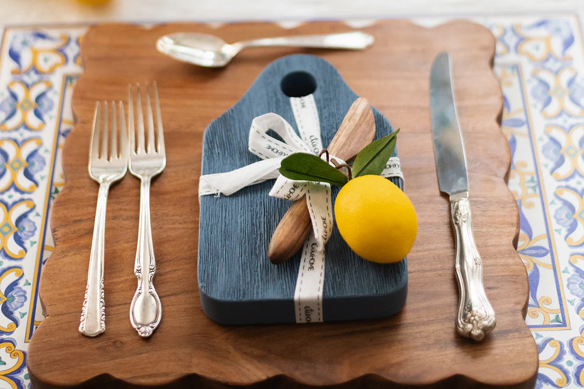 Decorative cutting board with a lemon, knife, and forks on a patterned tablecloth.