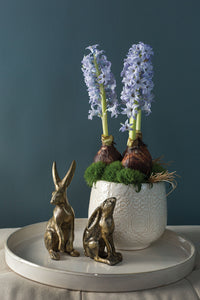 White ceramic pot with purple flowers and greenery, accompanied by two small bronze rabbit sculptures on a dark background.