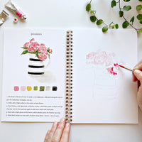 Open book with watercolor flowers and instructions with hand holding a paintbrush, on a white surface.