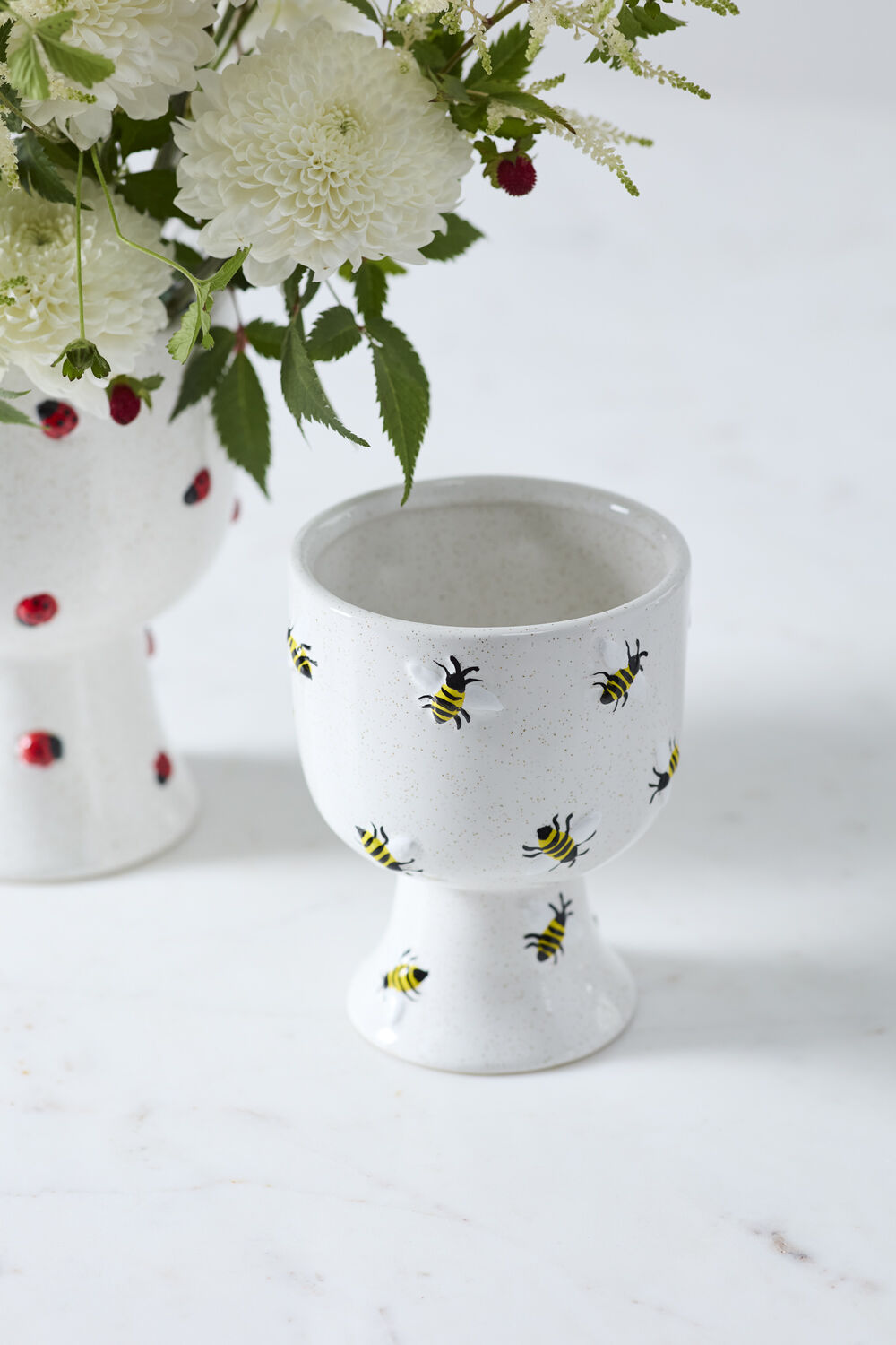 White ceramiccompote with bee design on a white surface with flowers in the background