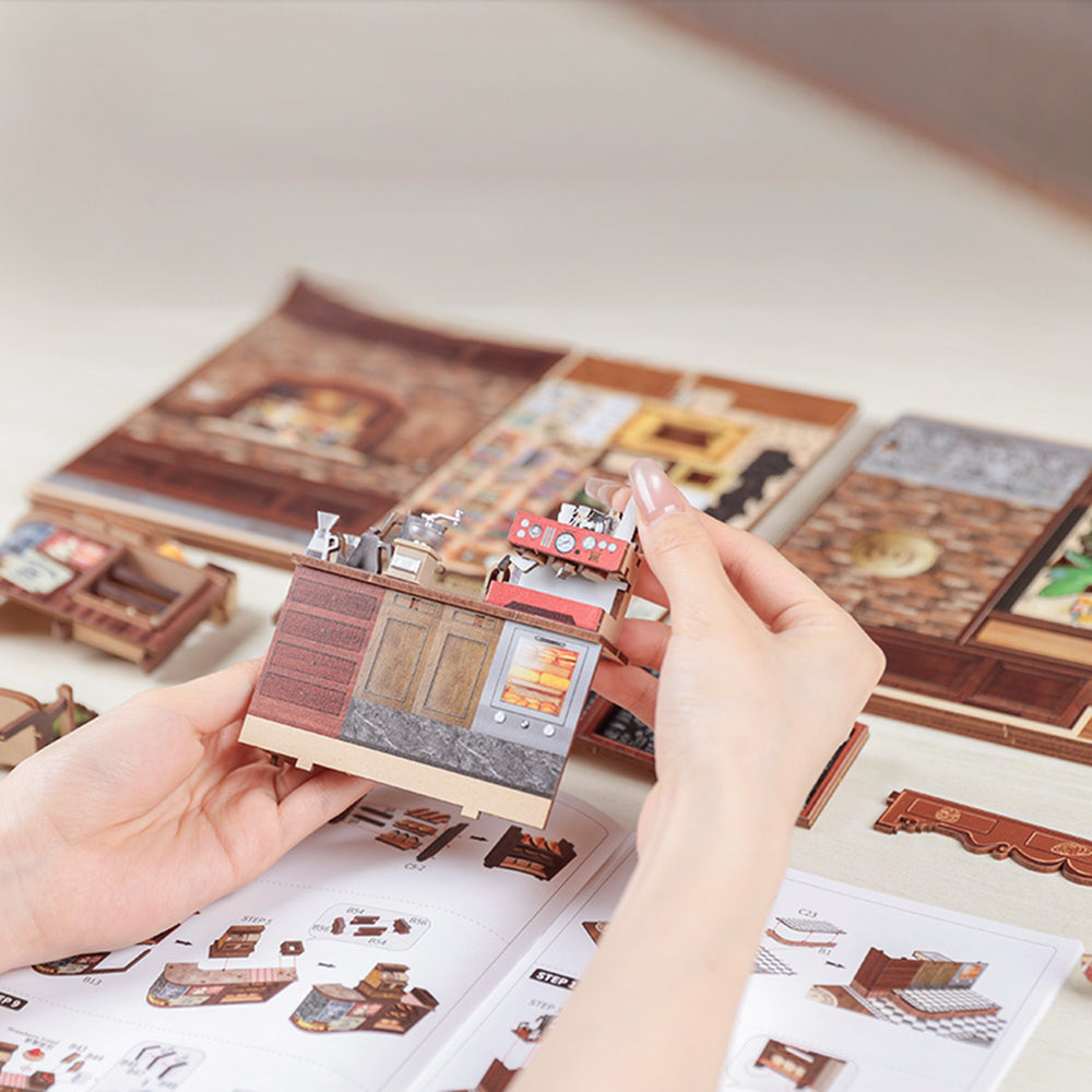 Hand holding a miniature model of a building with a detailed interior, against a background of similar models and a manual.