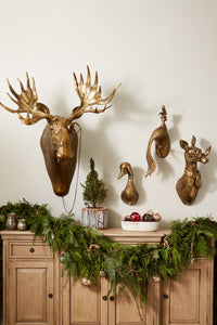 Decorative wall with gold moose head with monocle, duck with helmet, and other gold mounted animal busts above a wooden cabinet with greenery and small plants.