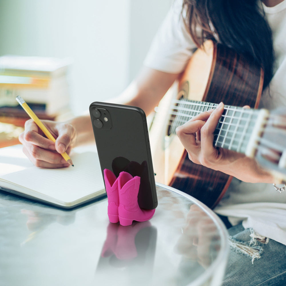 Person playing guitar and writing in a notebook with a pink phone holder on a glass table.