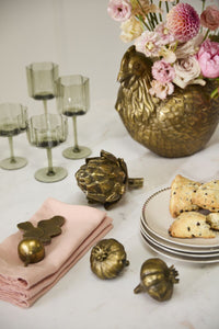 Decorative table setting with flowers, gilded vegetable figurines such as artichoke, radish, and garlic, next to pastries on plates on a white surface.