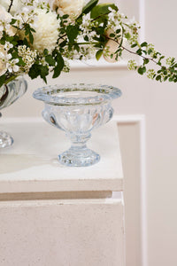 A clear glass pedestal bowl with a scalloped rim and footed base, styled on a white textured pedestal with white flowers in the background.