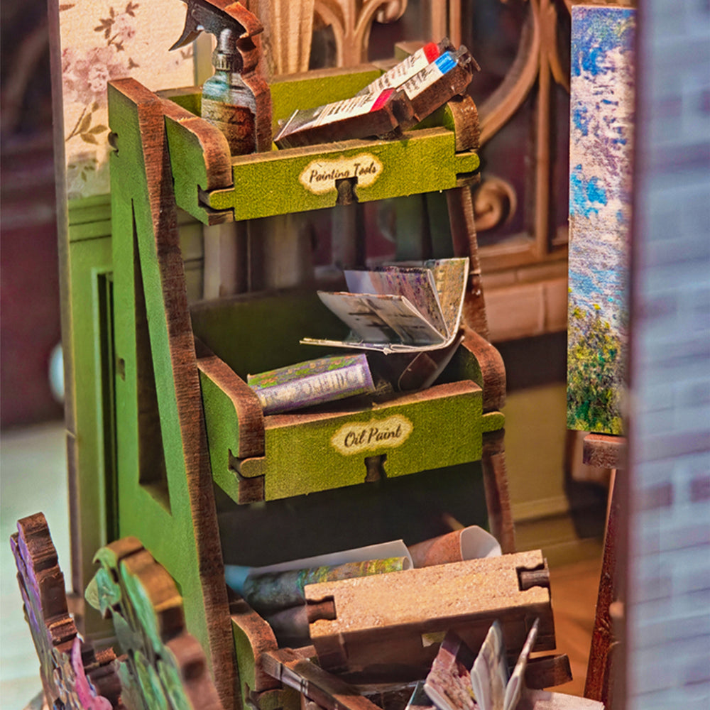 A miniature wooden artist's palette stand filled with tiny paint tubes, brushes, and art supplies, set against a blurred background of a workshop or studio.