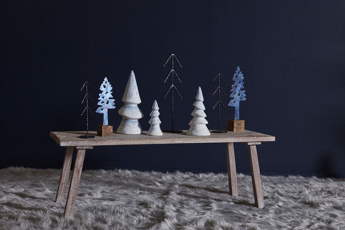 A rustic wooden table styled with a collection of decorative miniature Christmas trees in white, light blue, and black, set against a dark navy blue wall and a white shag rug.