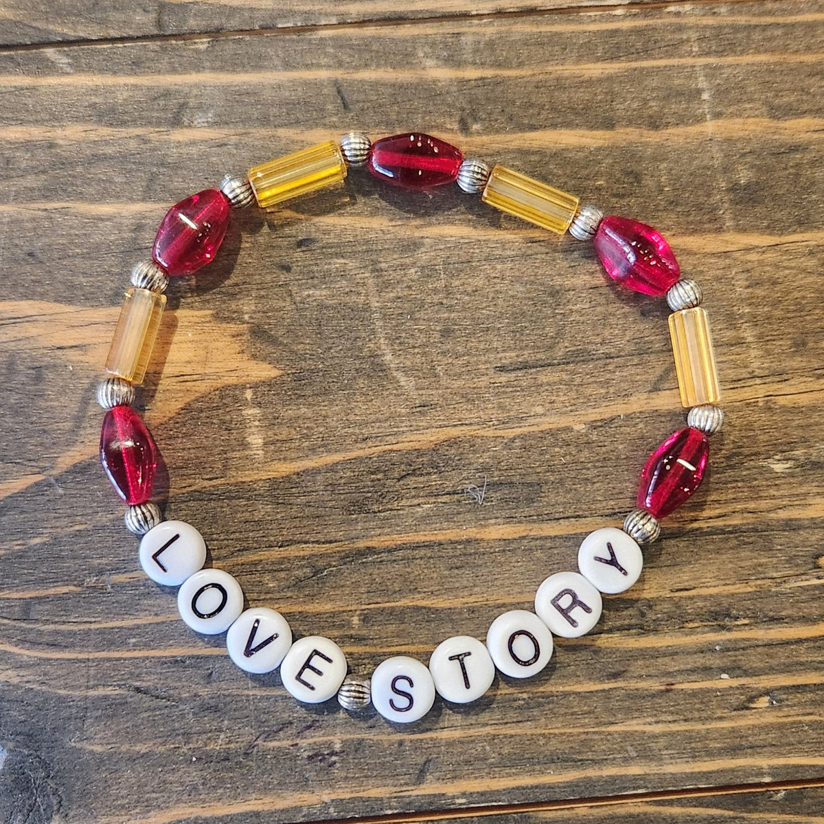 A beaded bracelet with faceted red beads, gold-toned cylindrical beads, and white letter beads spelling 'LOVE STORY' resting on a dark wood surface.