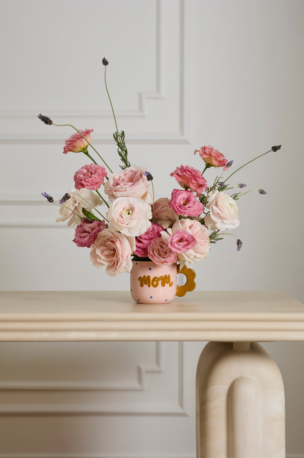 Bouquet of pink and white flowers in a mug labeled 'Mom' on a light wooden table.