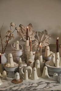 A Halloween decor display featuring ghost figurines and ghost candleholders with lit candles, in gray-white bowls and plates, with faux leaves and cotton, all on a marble surface.