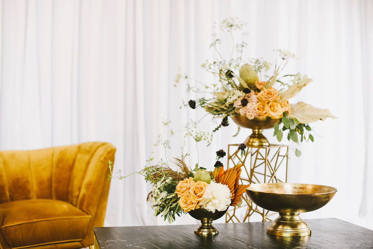 Decorative table setting with floral arrangements and gold bowls against a white curtain background.