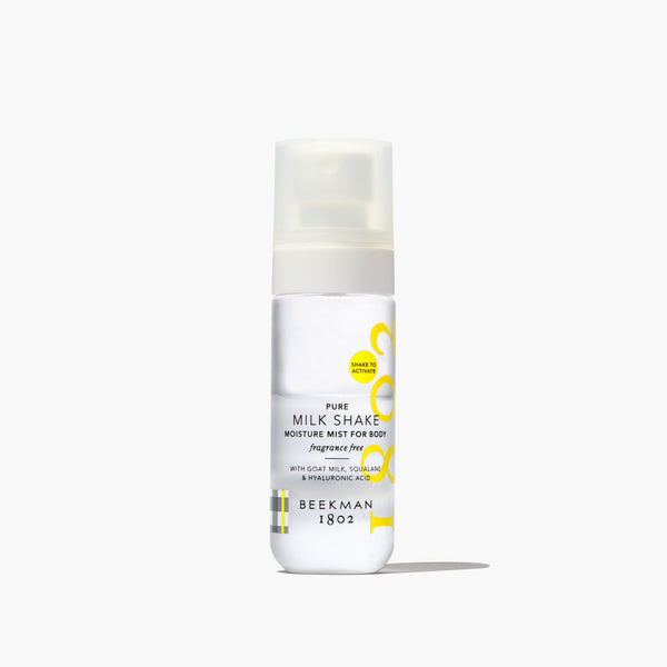 A white pump bottle of Beekman 1802 Pure Milk Shake Moisture Mist for Body, featuring a frosted cap and yellow graphic accents, set against a clean white background with a soft shadow.