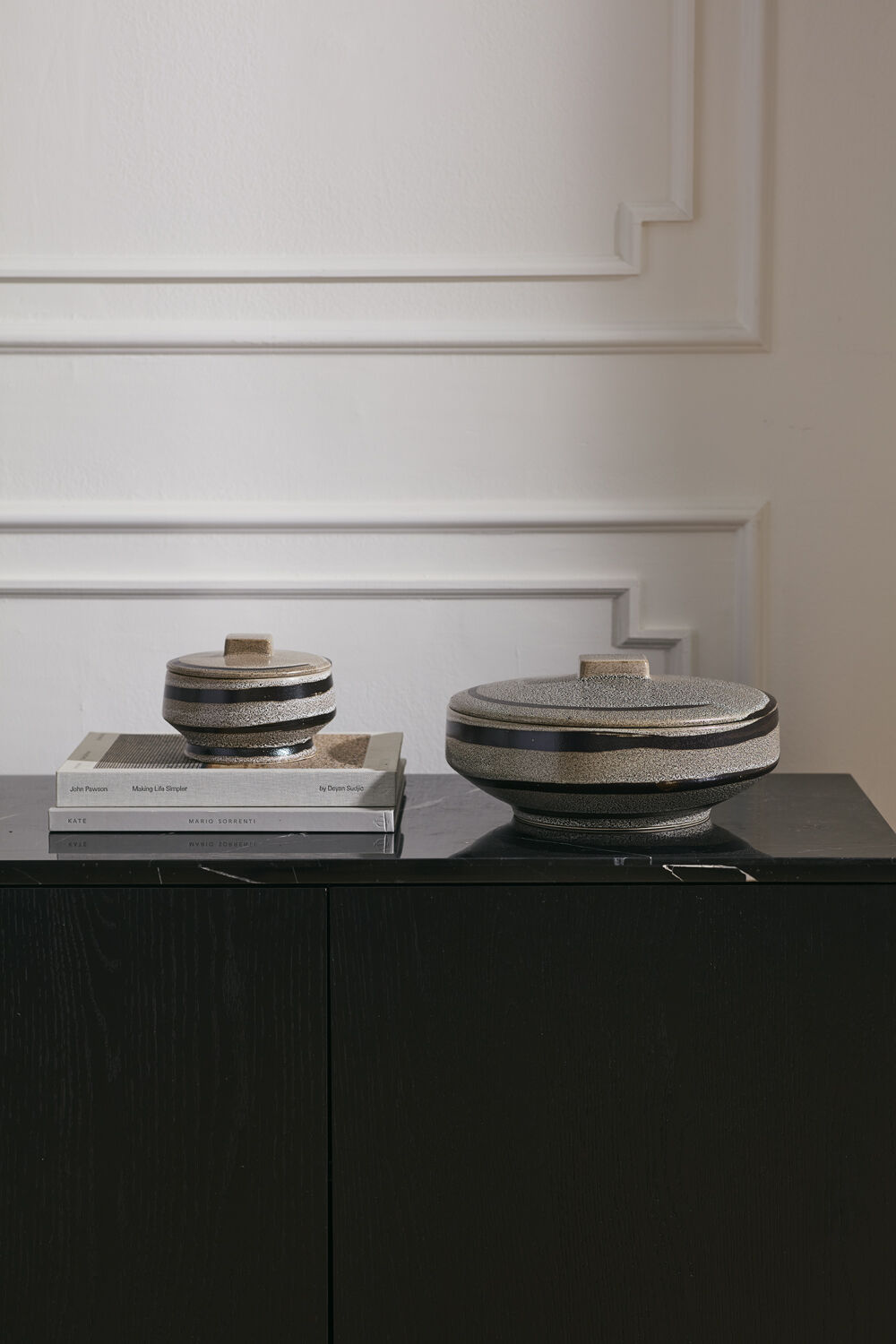 Two round lidded ceramic boxes with striped patterns in beige, gray, and black, displayed on a black sideboard against a white paneled wall.