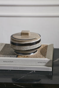 A round ceramic lidded box with a speckled beige and black glaze sits on top of two stacked books on a dark marble surface against a white paneled wall.