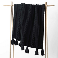 Black cable knit throw blanket with tassels on a wooden rack against a white background