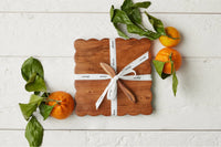 Wooden cheese board with a white ribbon, surrounded by oranges and leaves on a light wooden surface.
