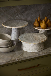 Speckled stone cake and serving stands on a kitchen counter with bowls and pears.