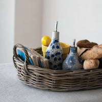 Wicker basket with kitchen items including blue ceramic oil cruets, yellow container, and brown loaf of bread on a light surface.