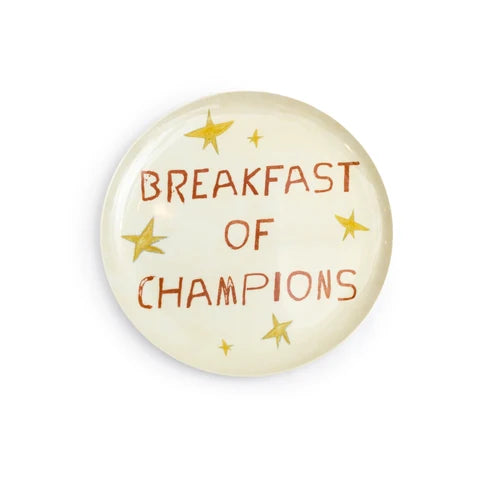 A round, glossy cream-colored melamine plate featuring the text 'BREAKFAST OF CHAMPIONS' in a rustic brown font, decorated with small gold stars, shown on a plain white background.