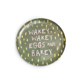 A round plate with a dark green glaze, decorated with yellow dotted border accents and the phrase 'WAKEY WAKEY EGGS AND BAKEY' in pink lettering, shown on a plain white background.