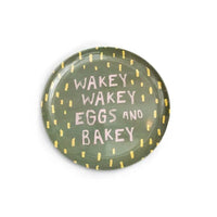 A round plate with a dark green glaze, decorated with yellow dotted border accents and the phrase 'WAKEY WAKEY EGGS AND BAKEY' in pink lettering, shown on a plain white background.