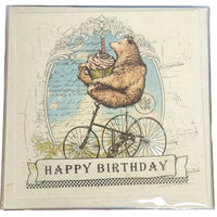 Birthday card with bear on a bicycle holding a cupcake, on a wooden surface.