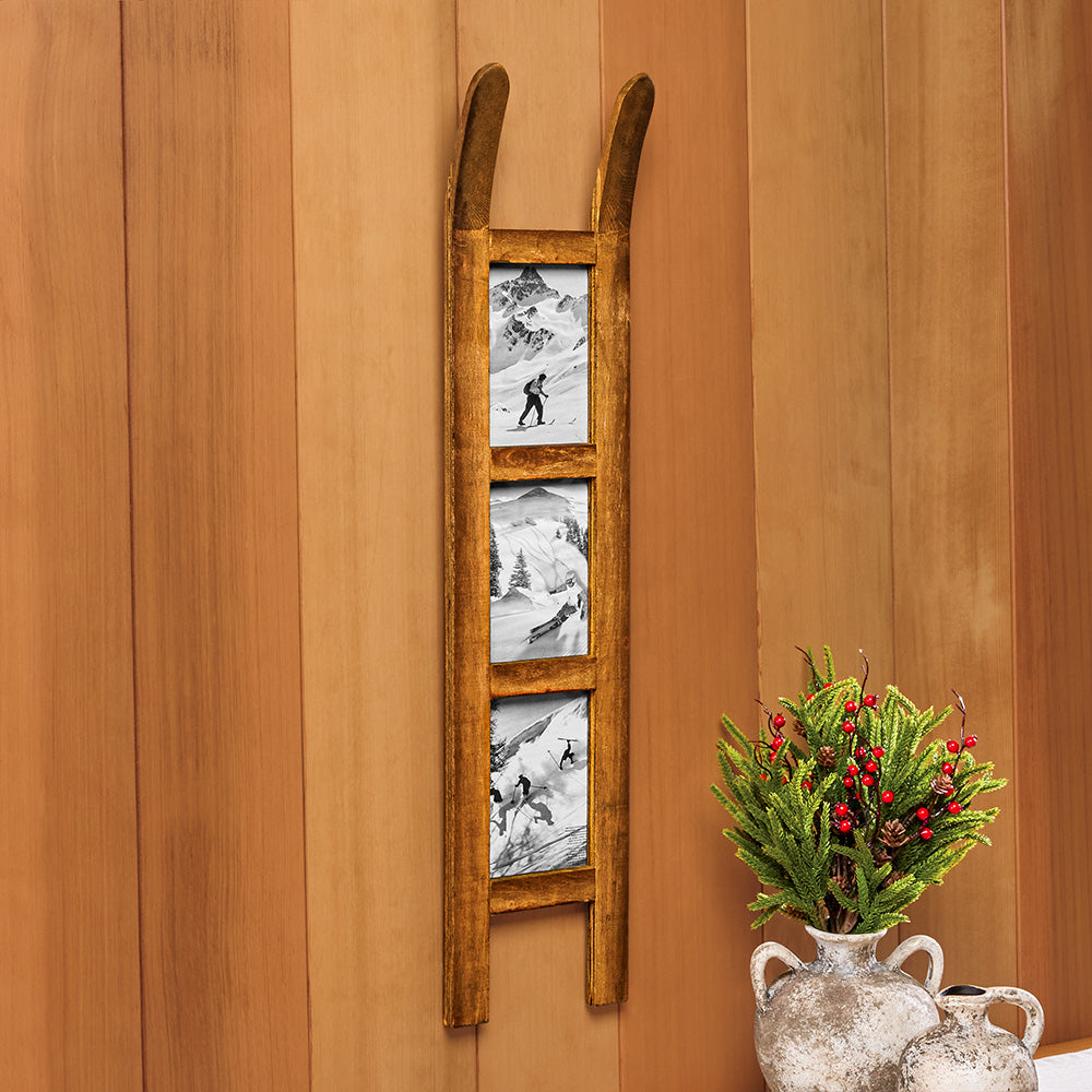 A rustic wooden ladder-shaped wall decor piece with three vertical panels showing black-and-white mountain and ski scenes, mounted on a warm wood-paneled wall beside a decorative vase with greenery and red berries.