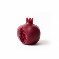 A sculptural candle shaped like a pomegranate, featuring a deep red rind with a textured, exposed seed cavity and a small white wick, on a white background.